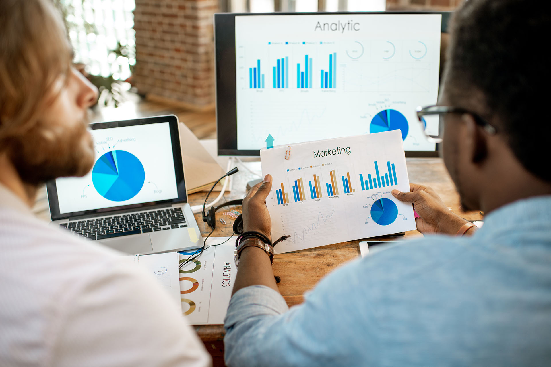 Two colleagues examining detailed marketing analytics charts with bar graphs and pie charts in an office, surrounded by laptops and reports.