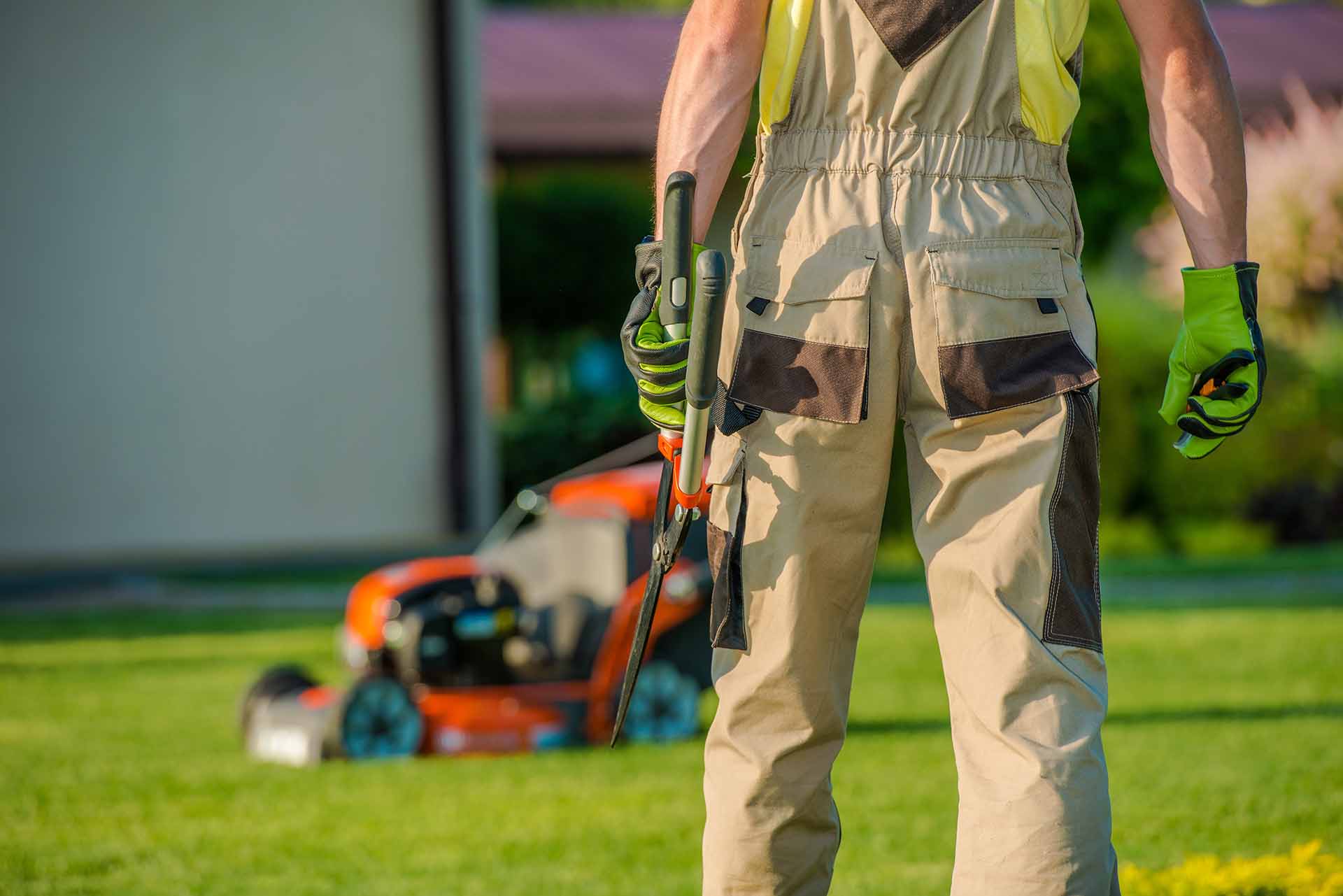 Gardener standing with tools in hand, ready to maintain a lawn, with a mower in the background.