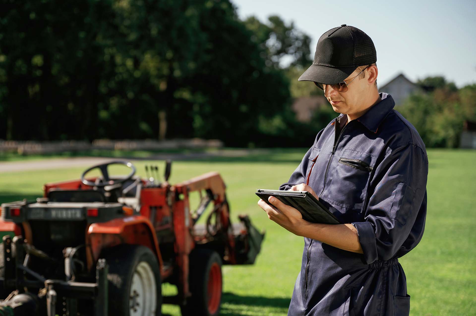 Man holding a tablet near a tractor, monitoring lawn care tasks and schedules.