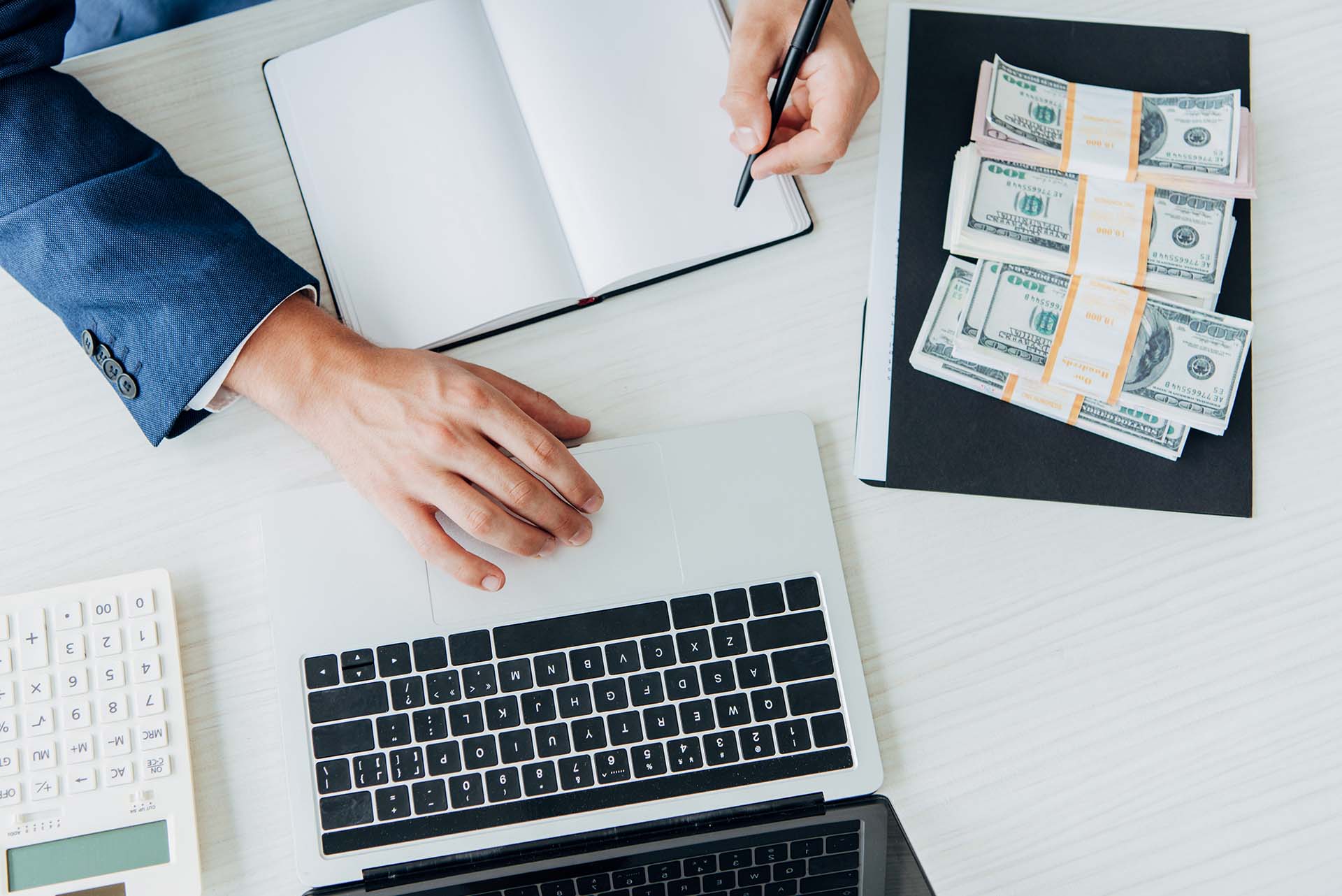 A businessman at a desk with a laptop, calculator, stacks of cash, and a notebook, writing financial plans.