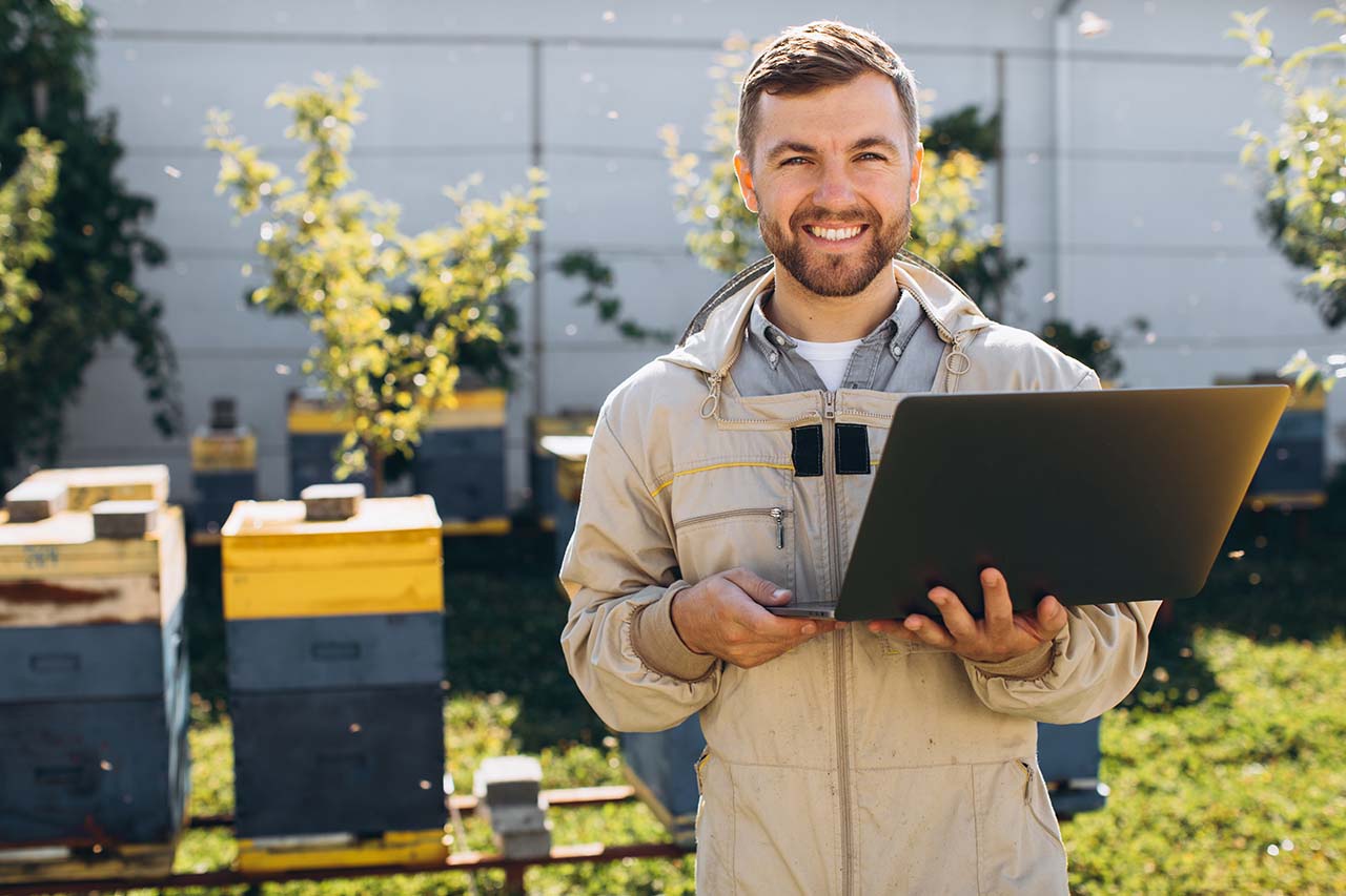 A smiling man in protective gear holding a laptop in a garden setting with beehives in the background.