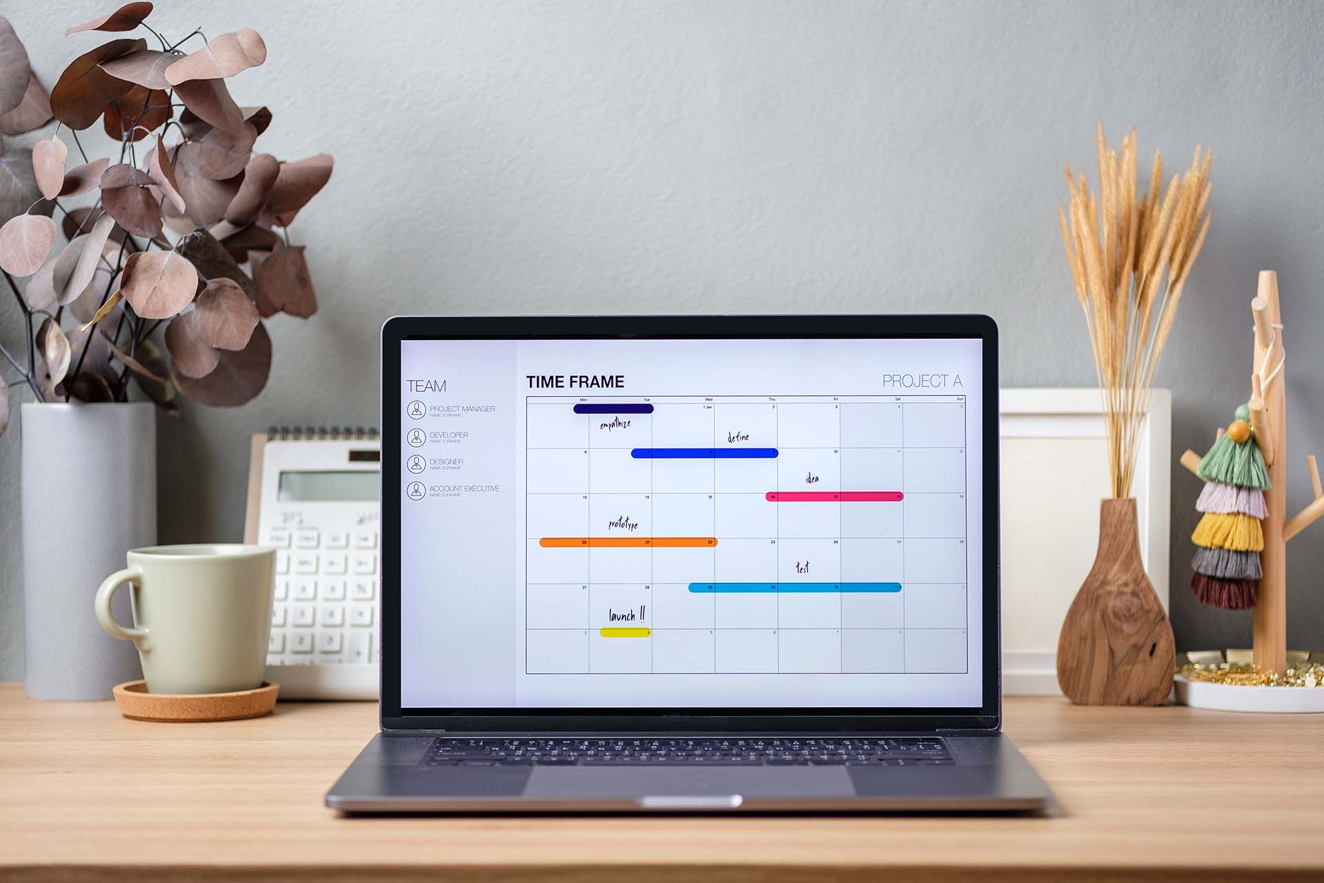 A laptop displaying a project timeline chart with tasks categorized by team roles, placed on a neat office desk with décor and a coffee mug.