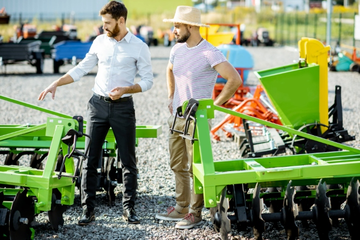 Landscaping equipment salesman discussing green machinery options with customer at outdoor dealership lot